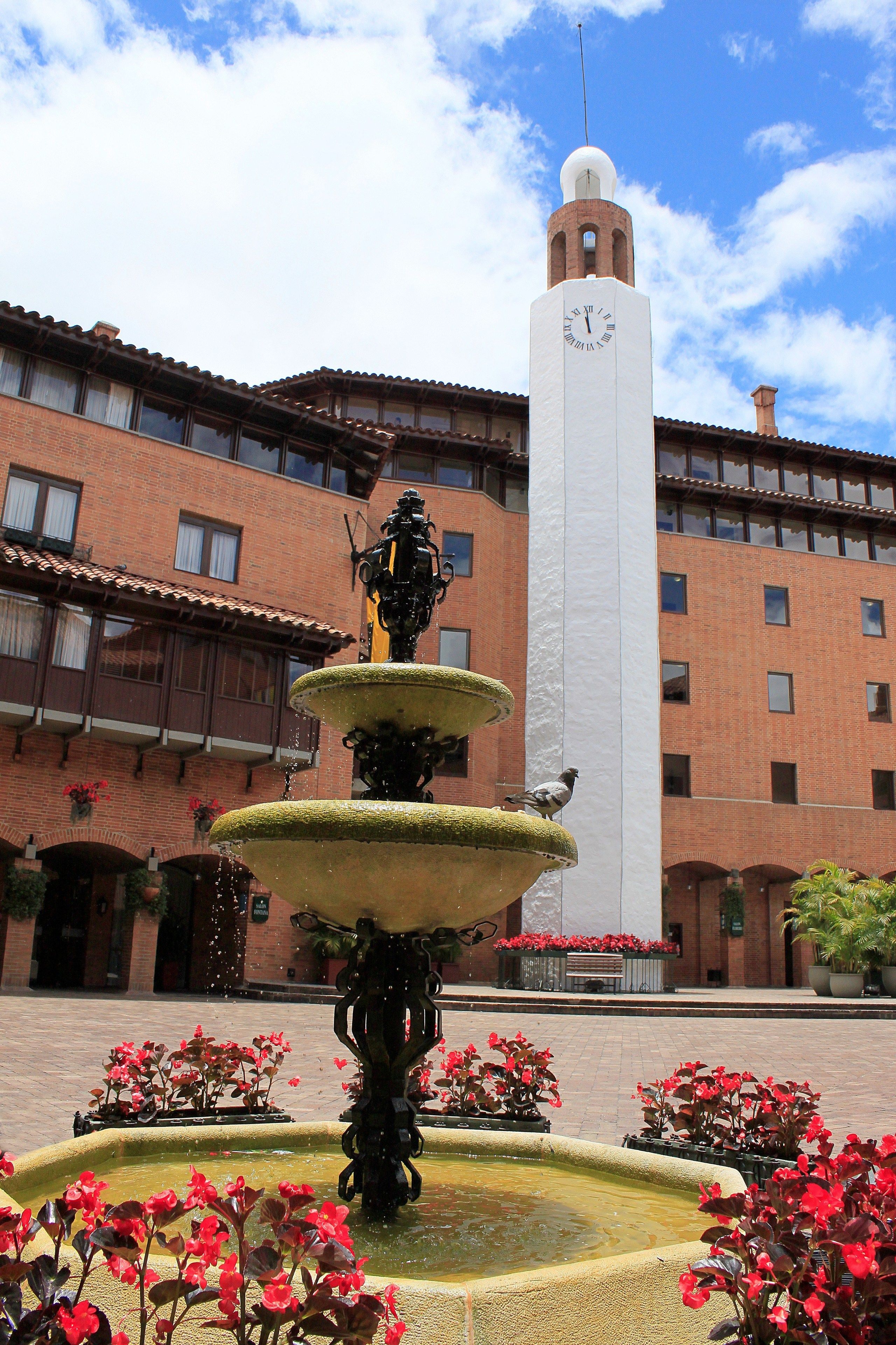 Un patio con una fuente de varios niveles, una torre del reloj y un edificio con muros de ladrillo rojo y balcones.