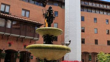 Un patio con una fuente de varios niveles, una torre del reloj y un edificio con muros de ladrillo rojo y balcones.