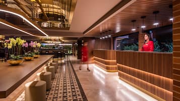 A modern hotel lobby with a long reception desk, a patterned floor, and a bar area with plants and bottles.