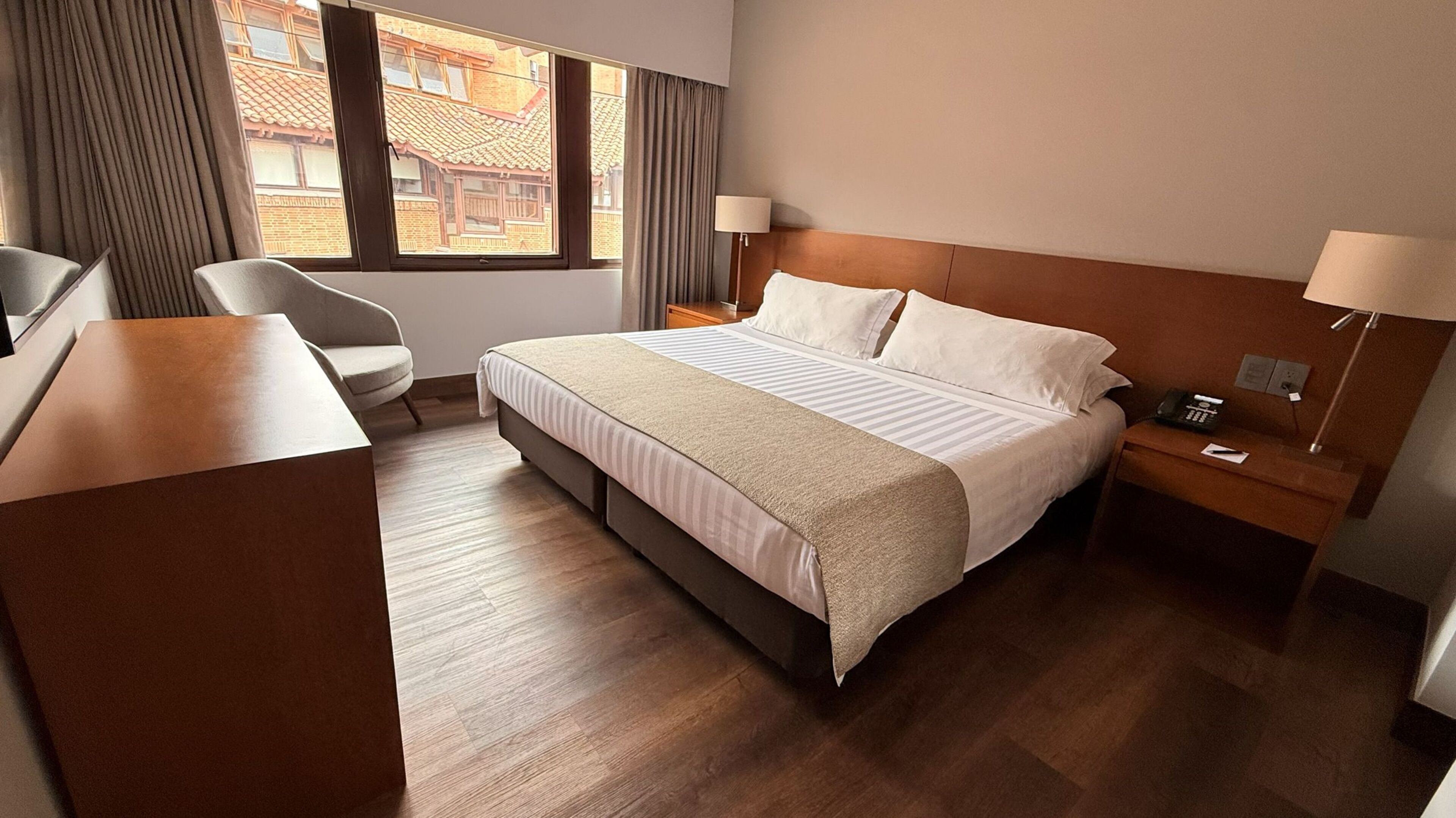 A hotel room with a bed, a desk, a chair, a nightstand, and a window with curtains.