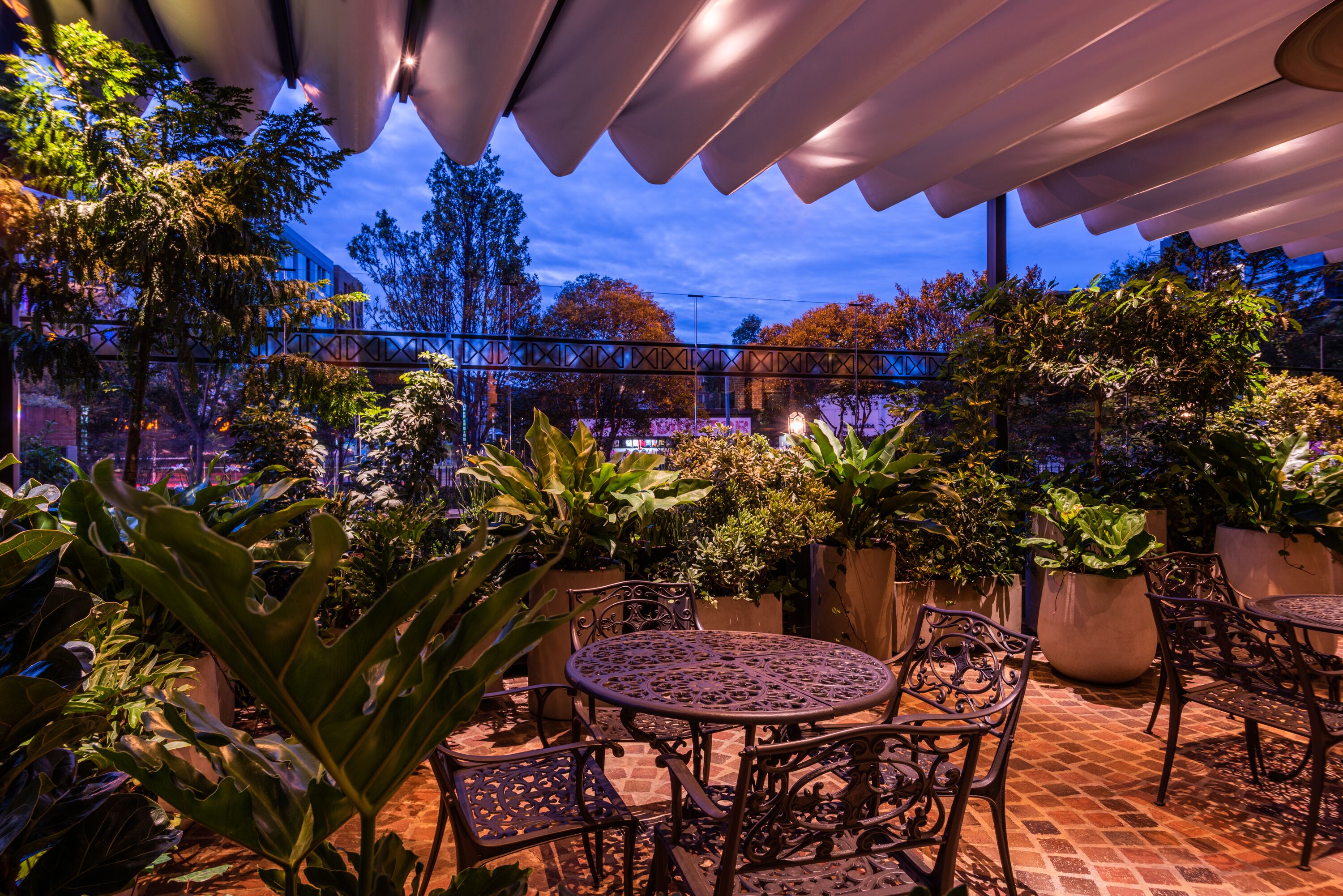 An outdoor patio with a round table and chairs, surrounded by potted plants and a metal fence.