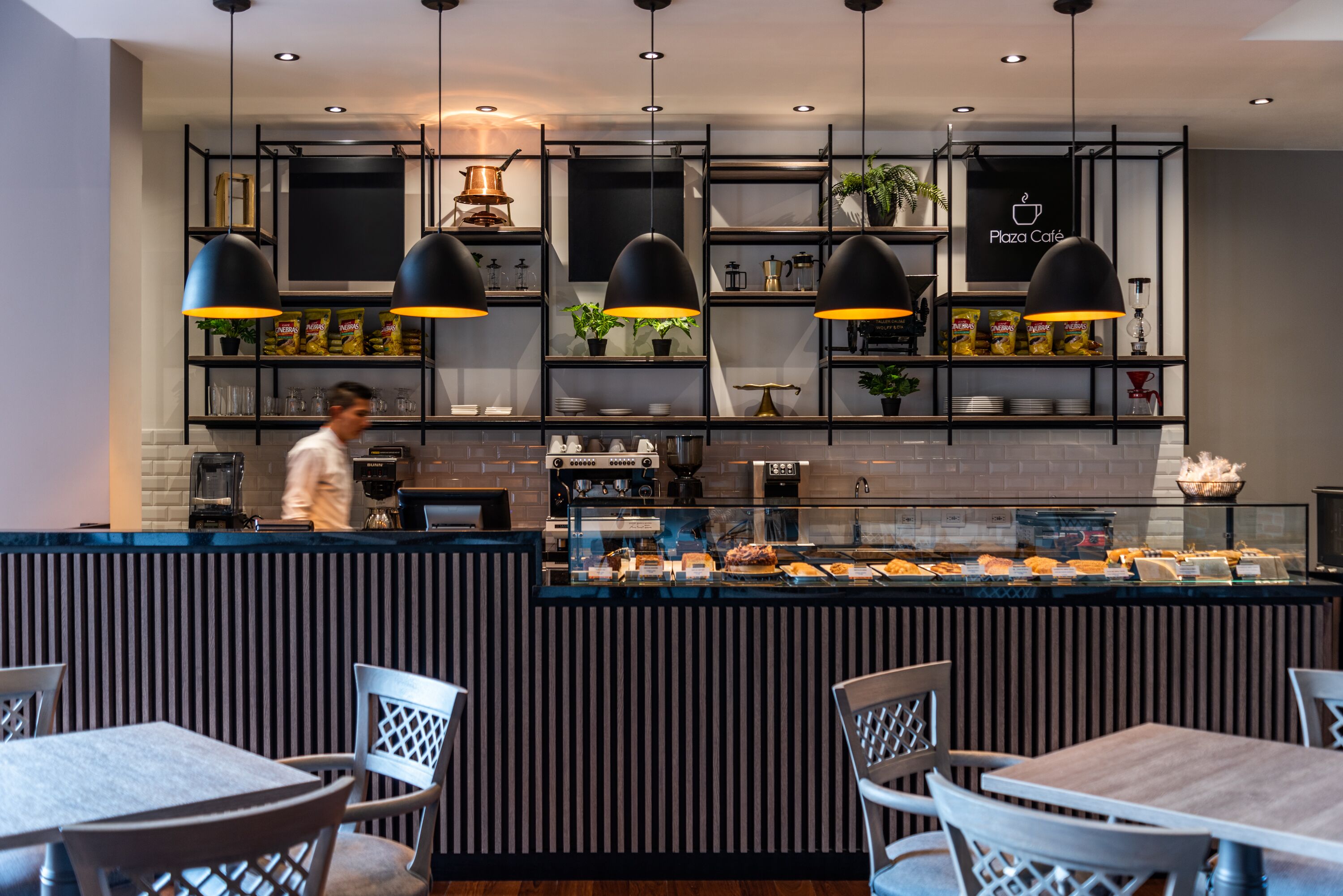 A modern café with a counter displaying various food items, black pendant lighting, and a person in the background.
