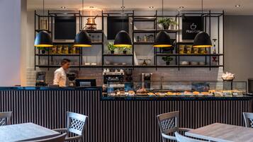 A modern café with a counter displaying various food items, black pendant lighting, and a person in the background.