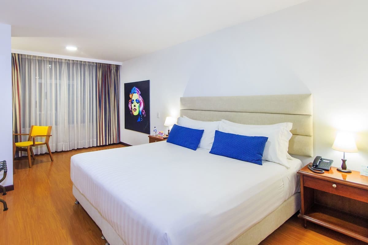 Ein Hotelzimmer mit einem großen Bett, einem Schreibtisch mit Lampe, einem Sessel und einem Fernseher an der Wand.