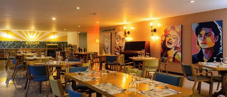 Un restaurante con varias mesas y sillas, una zona de cocina y una pared decorada con obras de arte coloridas.