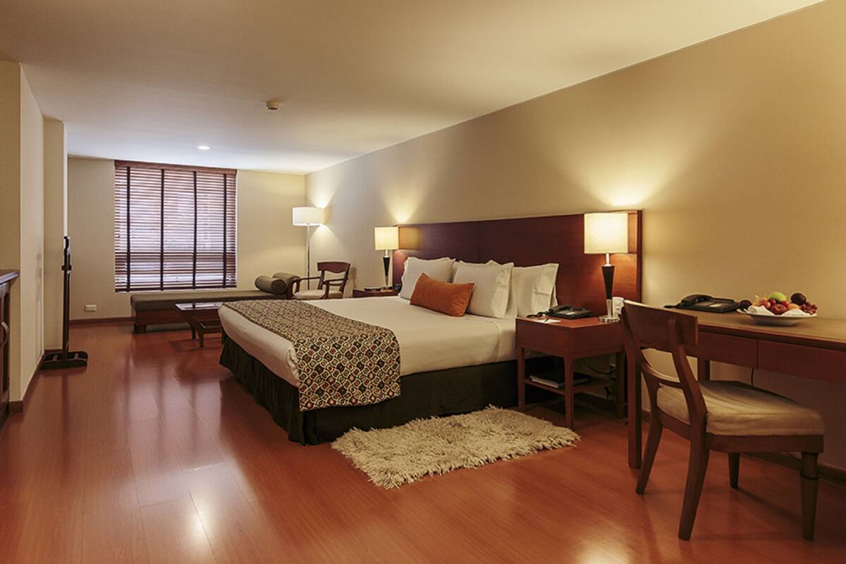Una habitación de hotel con cama, escritorio, silla, lámpara y ventana con persianas.