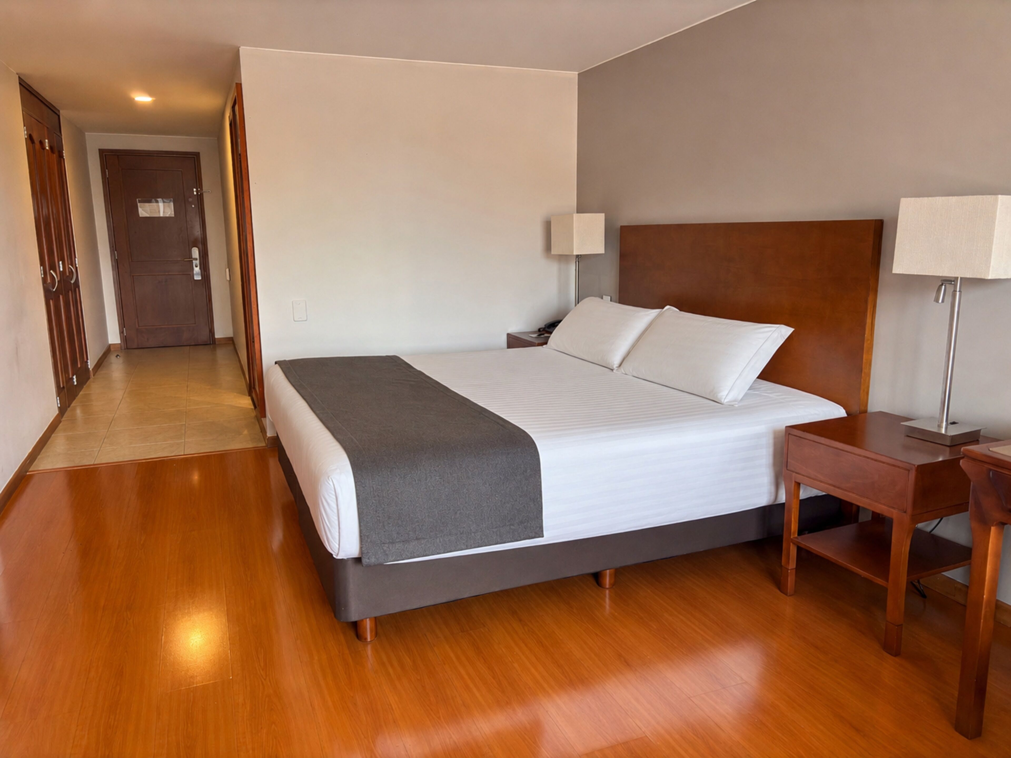 Quarto de hotel com cama, mesas de cabeceira, luminária e piso de madeira.