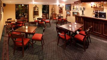 A restaurant with wooden tables and red upholstered chairs, a bar area with bottles, and framed artwork on the walls.