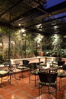 Un patio al aire libre con mesas y sillas de metal, rodeado de follaje y un suelo de baldosas.