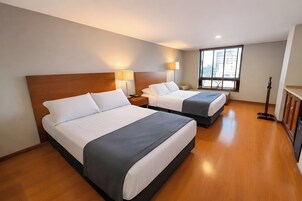Habitación de hotel con dos camas, una ventana con persianas, una mesita de noche con lámpara y suelo de madera.