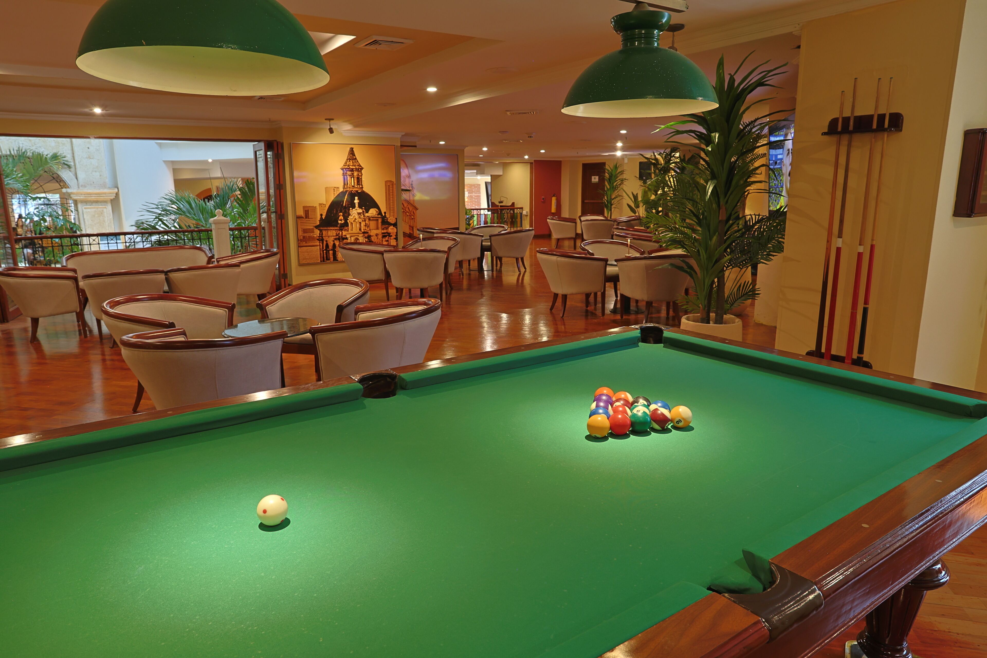 A pool table with balls, chairs, and a large green pendant light in a room with a view of a building and plants.