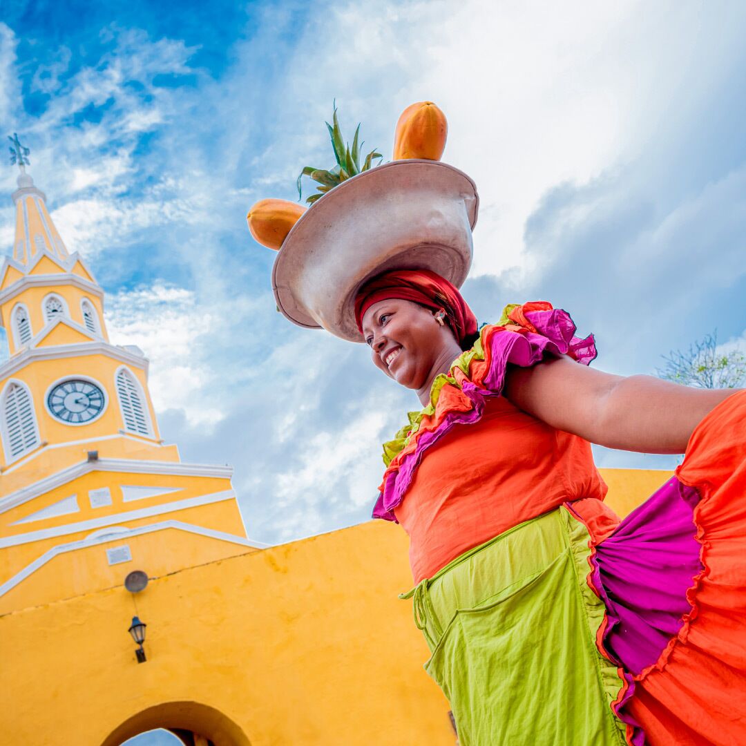 A person in a colorful costume with a large hat, standing in front of a yellow building with a clock tower.