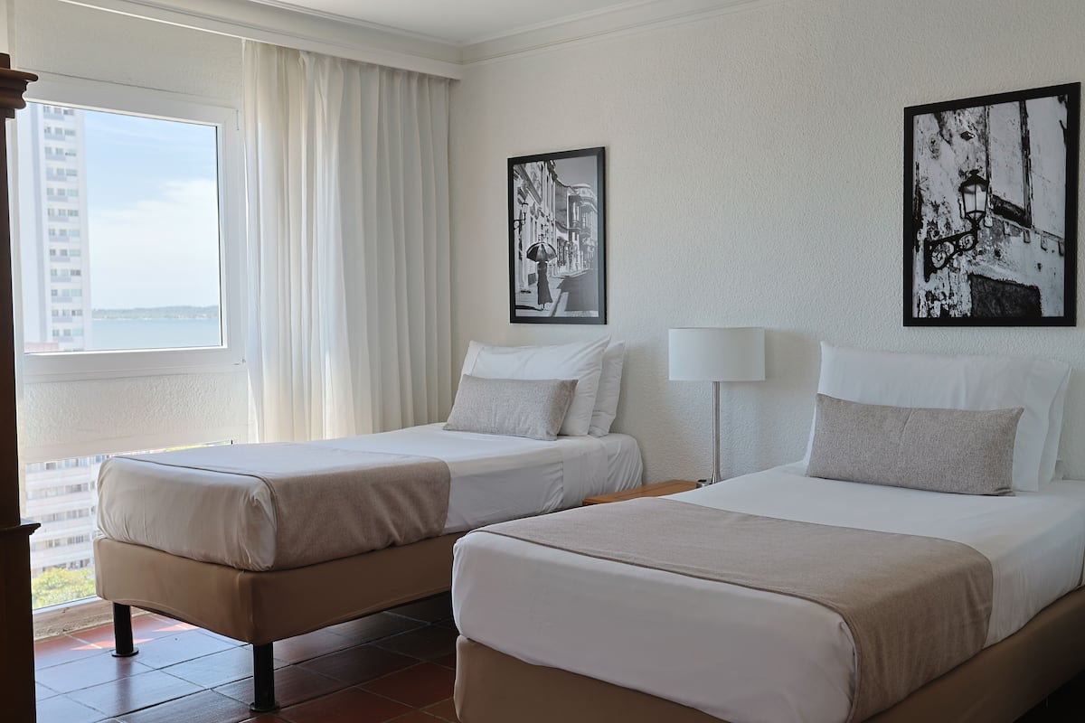 Een hotelkamer met twee bedden, een groot raam met gordijnen en twee zwart-wit foto’s aan de muur.