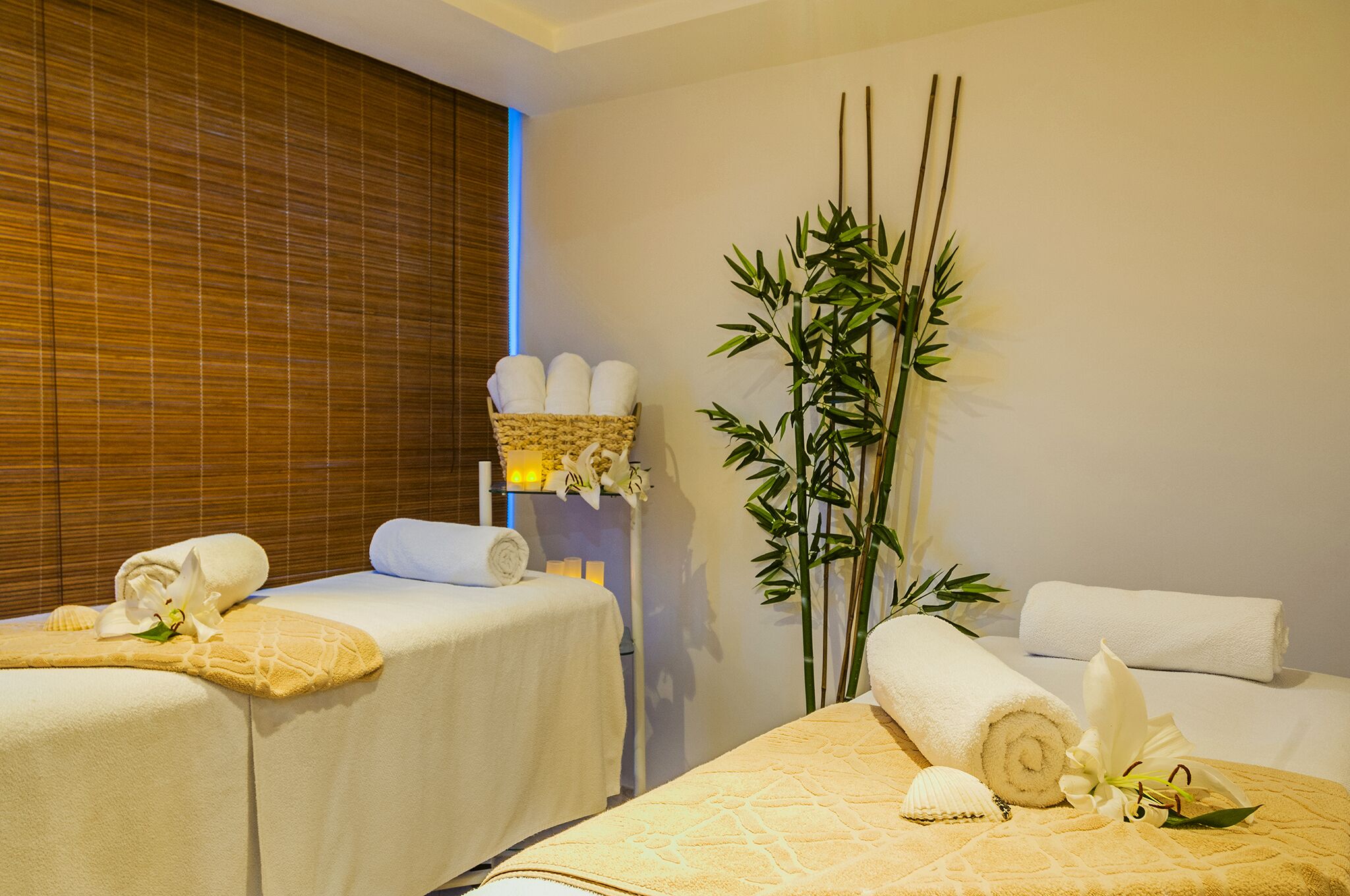 Un cuarto de spa con dos camillas de masaje, cada una con una toalla enrollada y un adorno. Se observa una planta de bambú y una orquídea blanca en la pared.