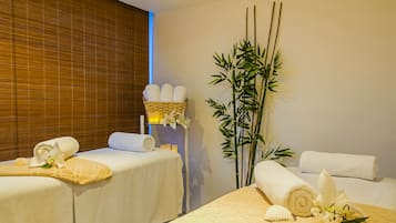 Un cuarto de spa con dos camillas de masaje, cada una con una toalla enrollada y un adorno. Se observa una planta de bambú y una orquídea blanca en la pared.