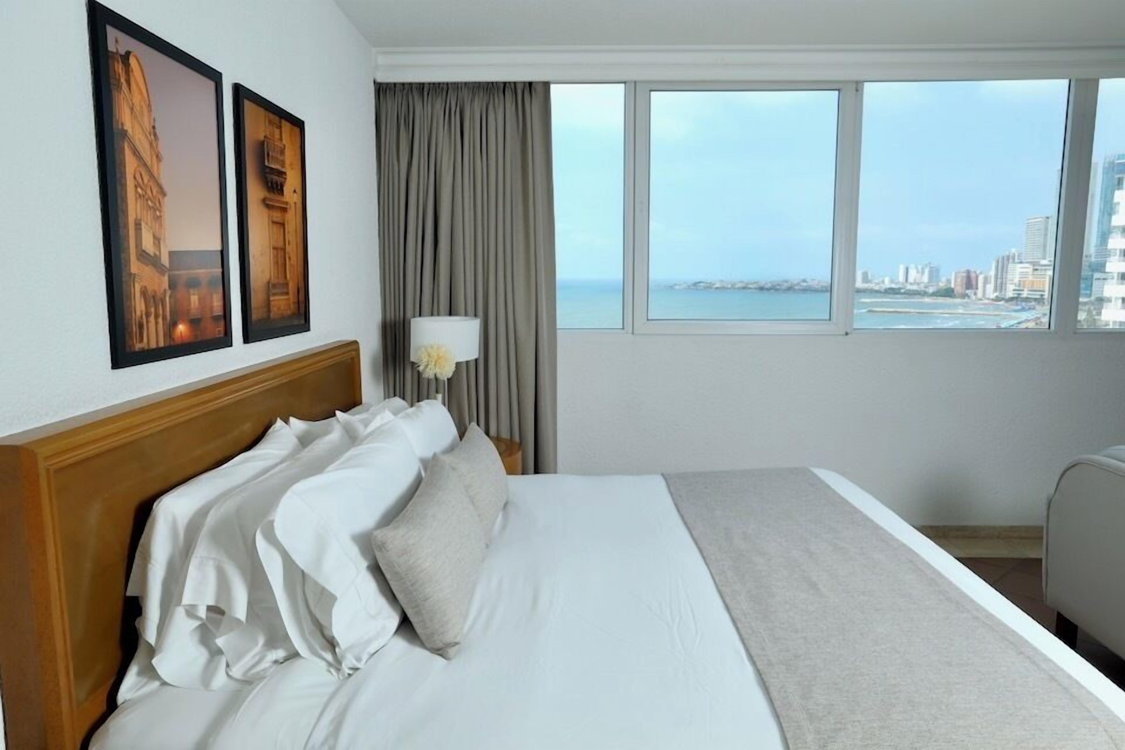 A hotel room with a large bed, two framed pictures on the wall, a window with a city view, and a lamp.