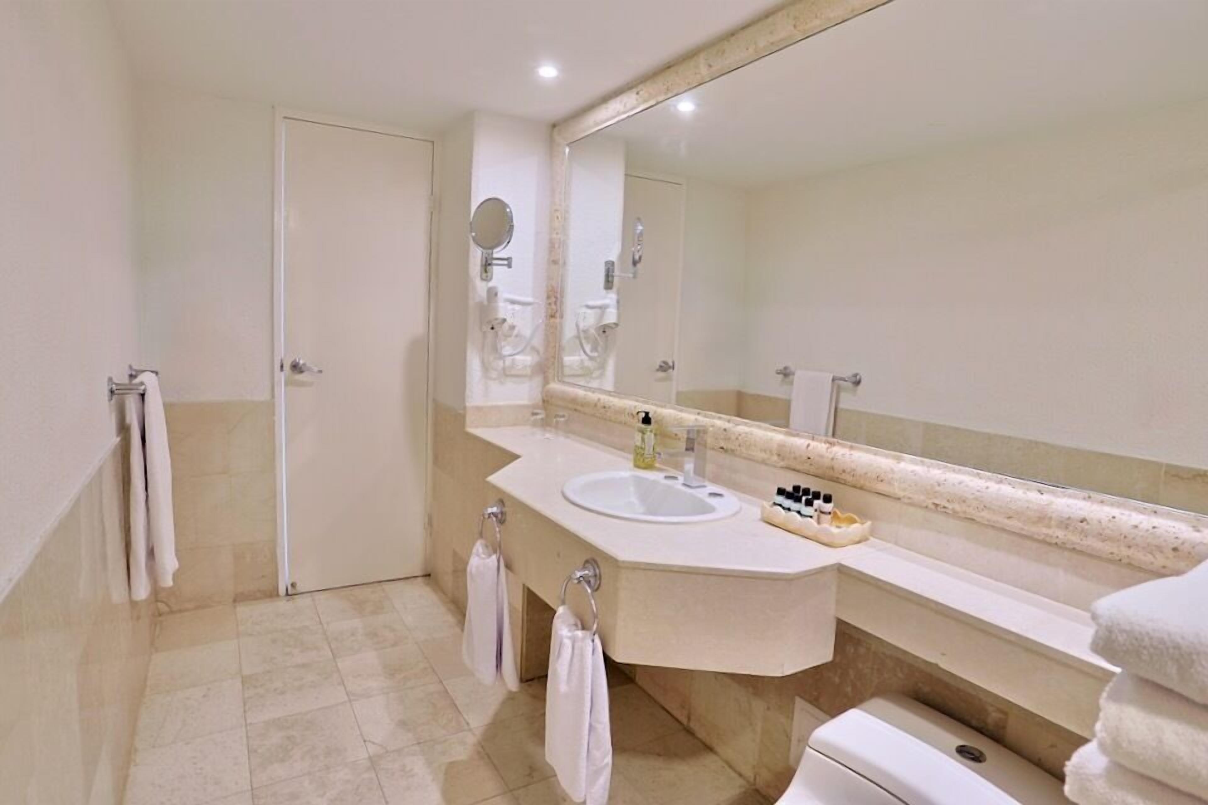 A bathroom with a white sink, a large mirror, and a toilet. There are towels hanging on the wall and a small shelf with toiletries.