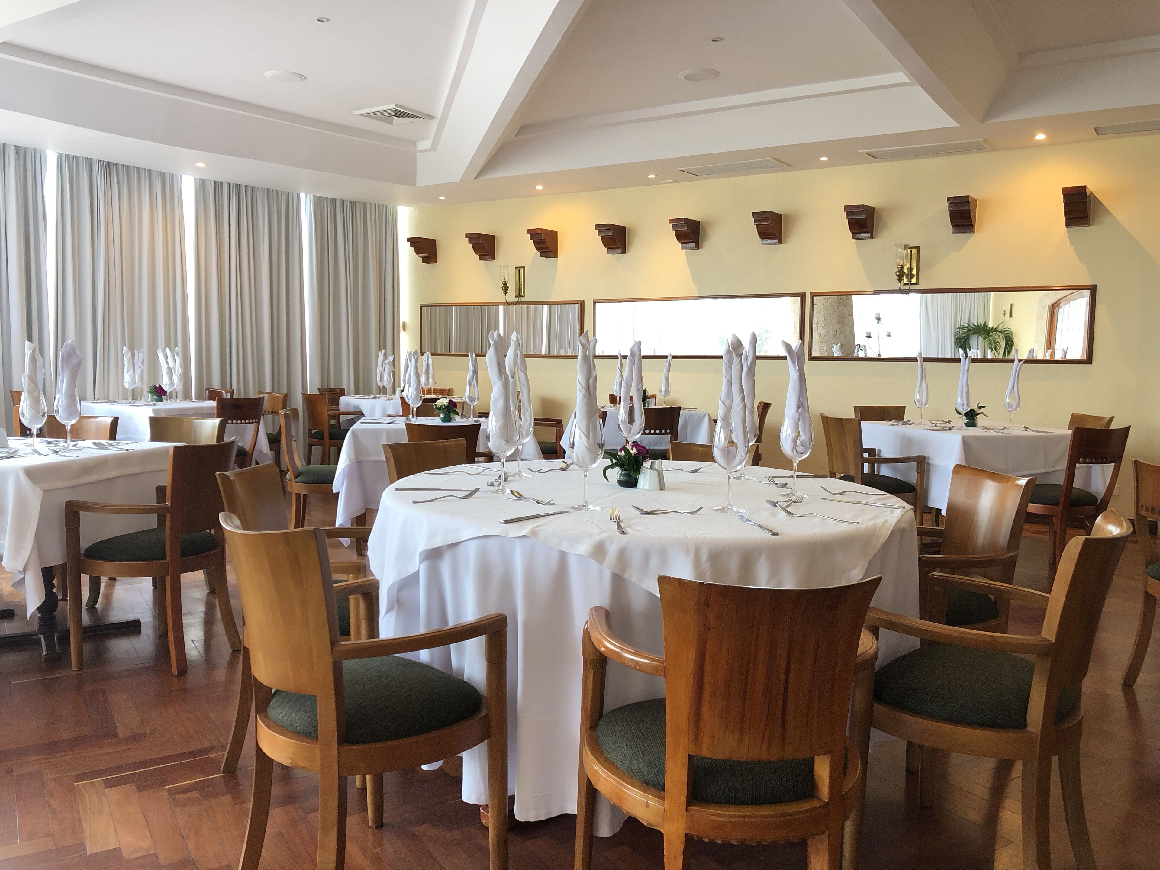 Uma sala de jantar com mesas redondas, cadeiras de madeira e toalhas de mesa brancas.