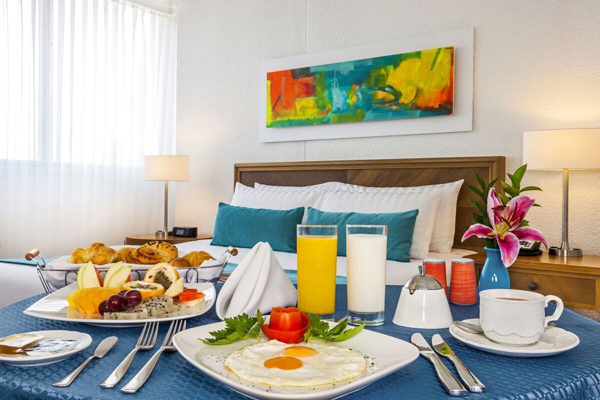 Ein Hotelzimmer mit Frühstückstisch, gedeckt mit Croissants, Obst, Eiern und Getränken, einem Bett mit blauen Kissen und einem farbenfrohen, abstrakten Gemälde an der Wand.