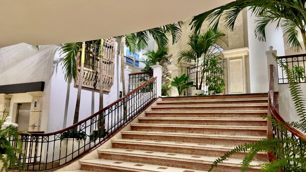 Una imponente escalera de mármol con barandillas de hierro forjado, rodeada de exuberante vegetación y arquitectura clásica.