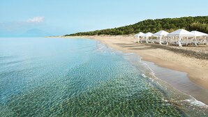 Una playa con agua cristalina, carpas blancas y una colina boscosa al fondo.