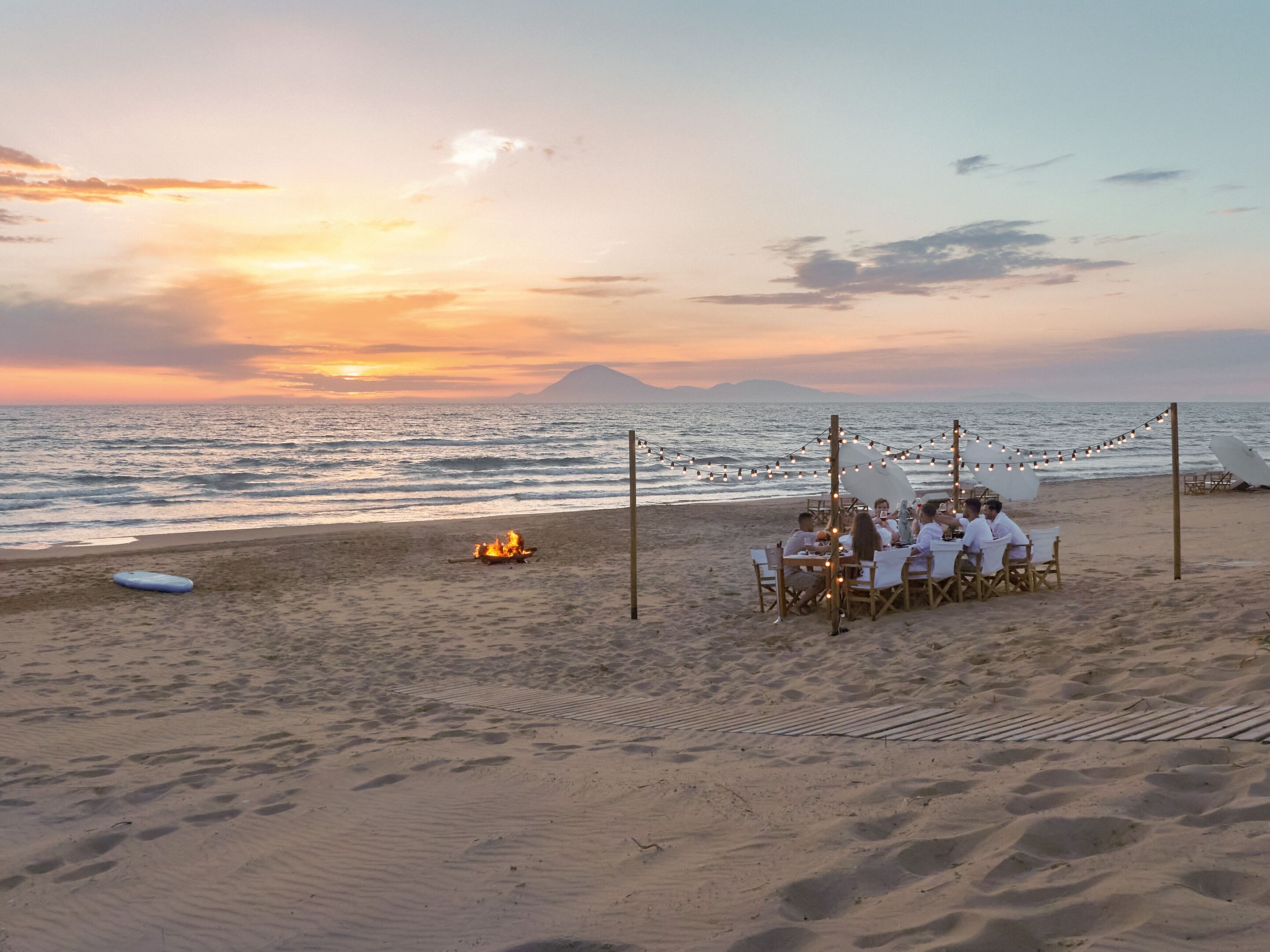 Montaje para comer frente al mar con una mesa y sillas bajo luces colgantes, con vista al océano y el atardecer de fondo.