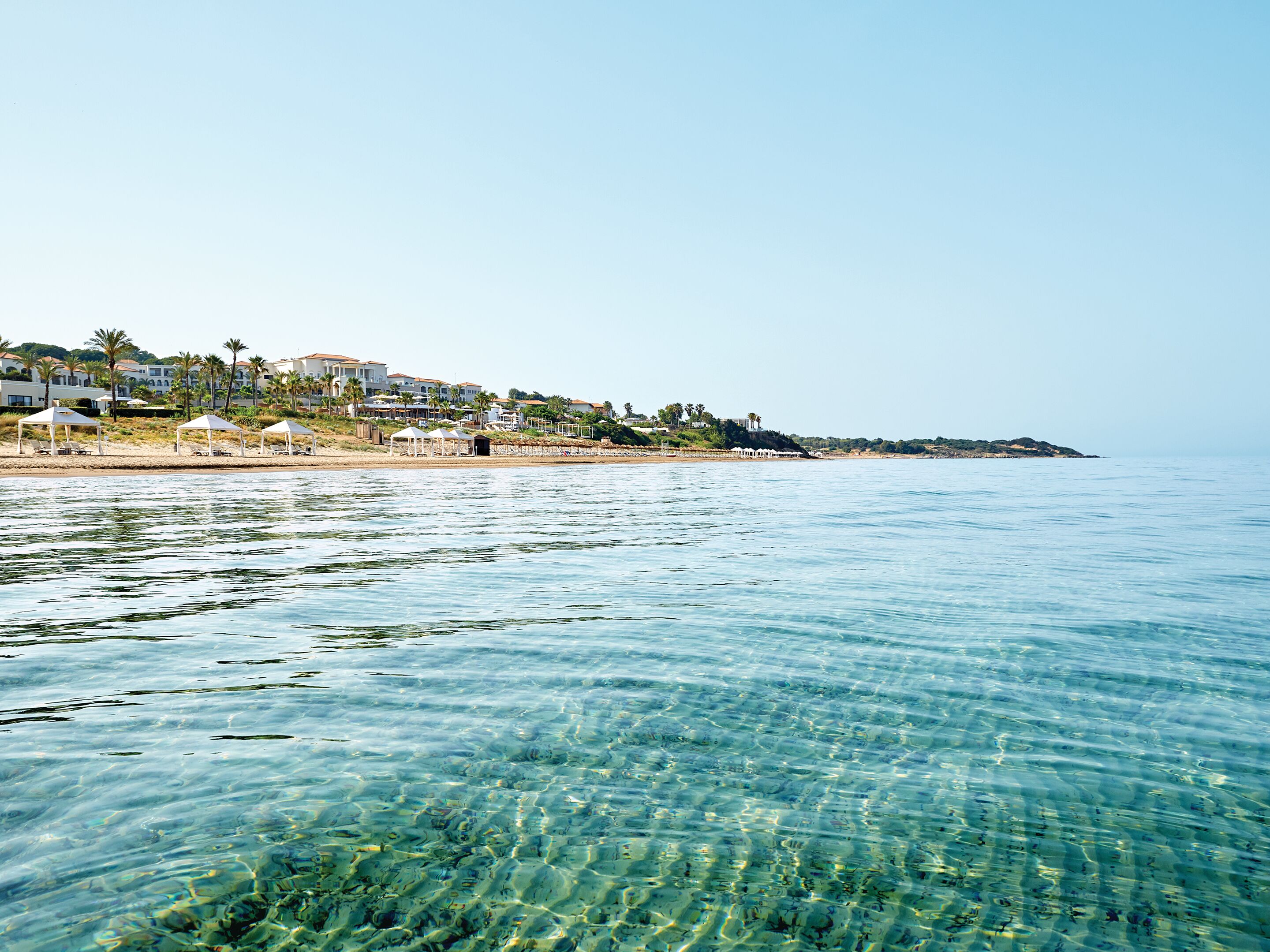 A coastal area with clear water, beach umbrellas, and buildings in the background.