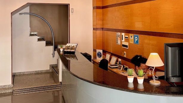 A reception area with a curved counter, multiple wicker pendant lights, a reception desk, a television, and a few potted plants. - Hotel Piccadilly Sitges (Sitges)