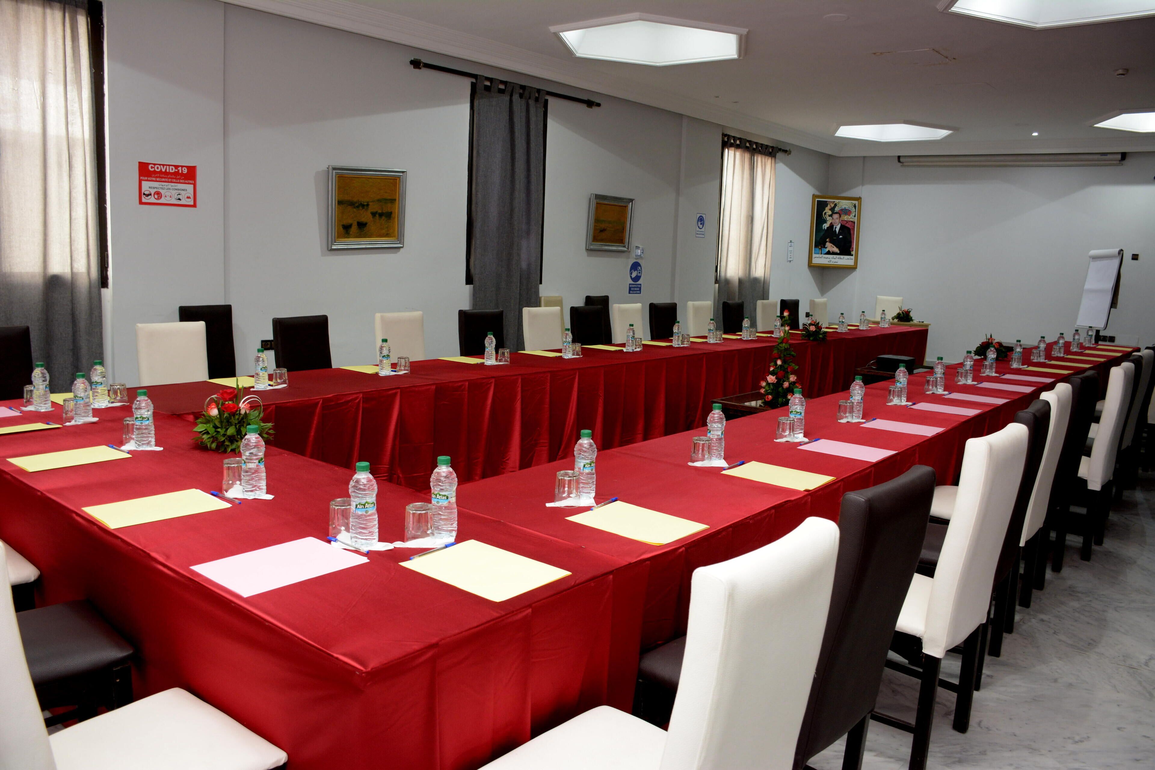 Un salón de conferencias con una larga mesa, sillas, botellas de agua y un mantel rojo.