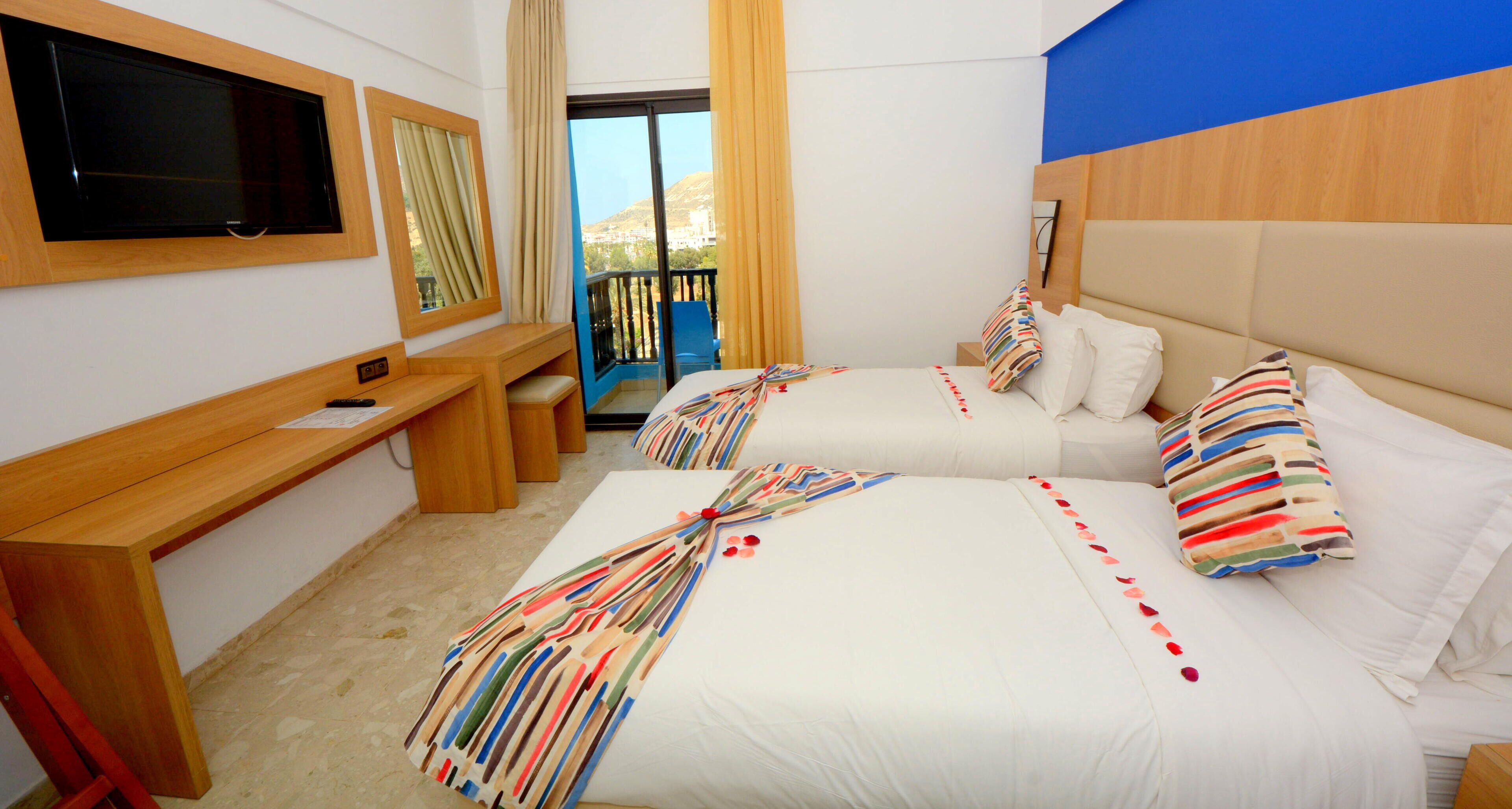 A hotel room with two beds, a desk, a TV, and a balcony with a view.