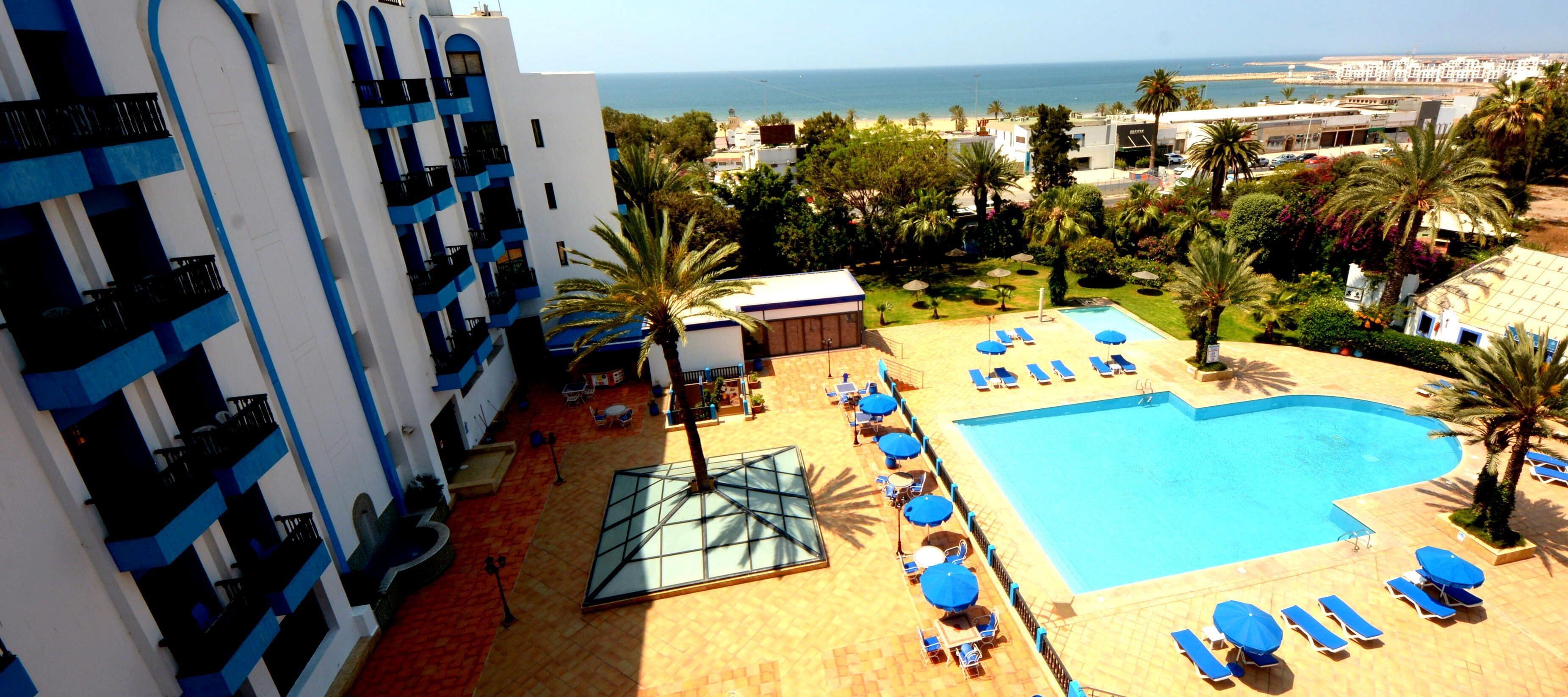 A hotel with a pool area, including sun loungers and umbrellas, surrounded by palm trees and a view of the sea.