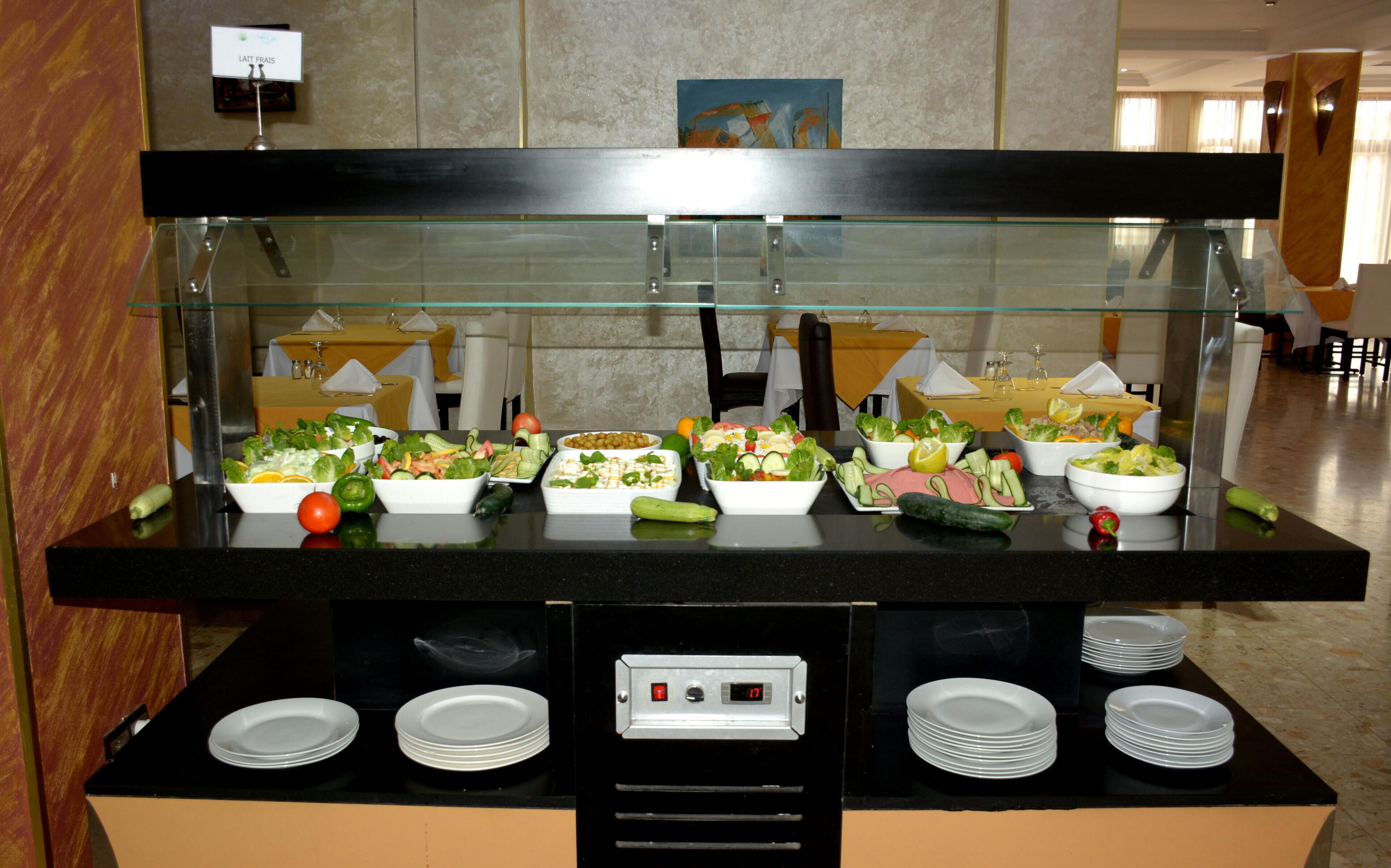 A buffet with various salads and fruits, a digital clock, and neatly stacked plates.