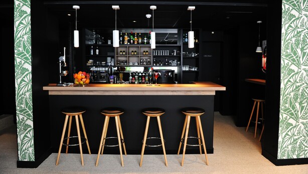 A bar area with a wooden counter, three stools, and shelves stocked with bottles and glasses. - Urban Style Hotel de France (Vannes)