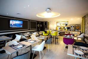A modern restaurant with a TV screen displaying a news program, multiple dining tables with chairs, and a kitchen area in the background.