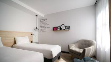 Ein Hotelzimmer mit zwei Betten, einem Sessel, einem kleinen Tisch sowie einer Wand mit gerahmten Bildern und einem motivierenden Zitat.