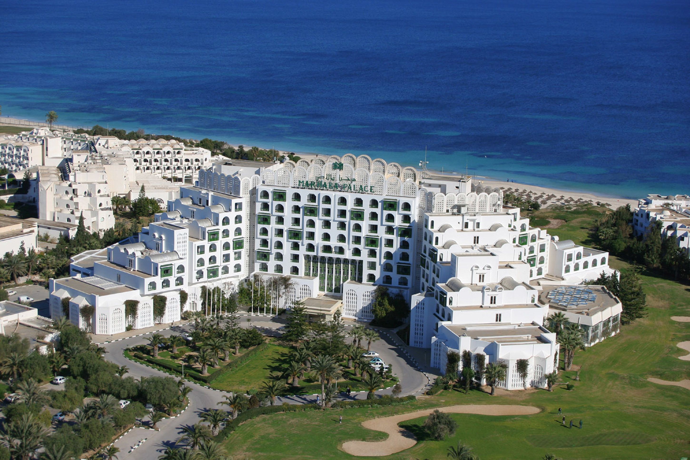 A large, white, multi-story hotel with a central courtyard and a beach view.
