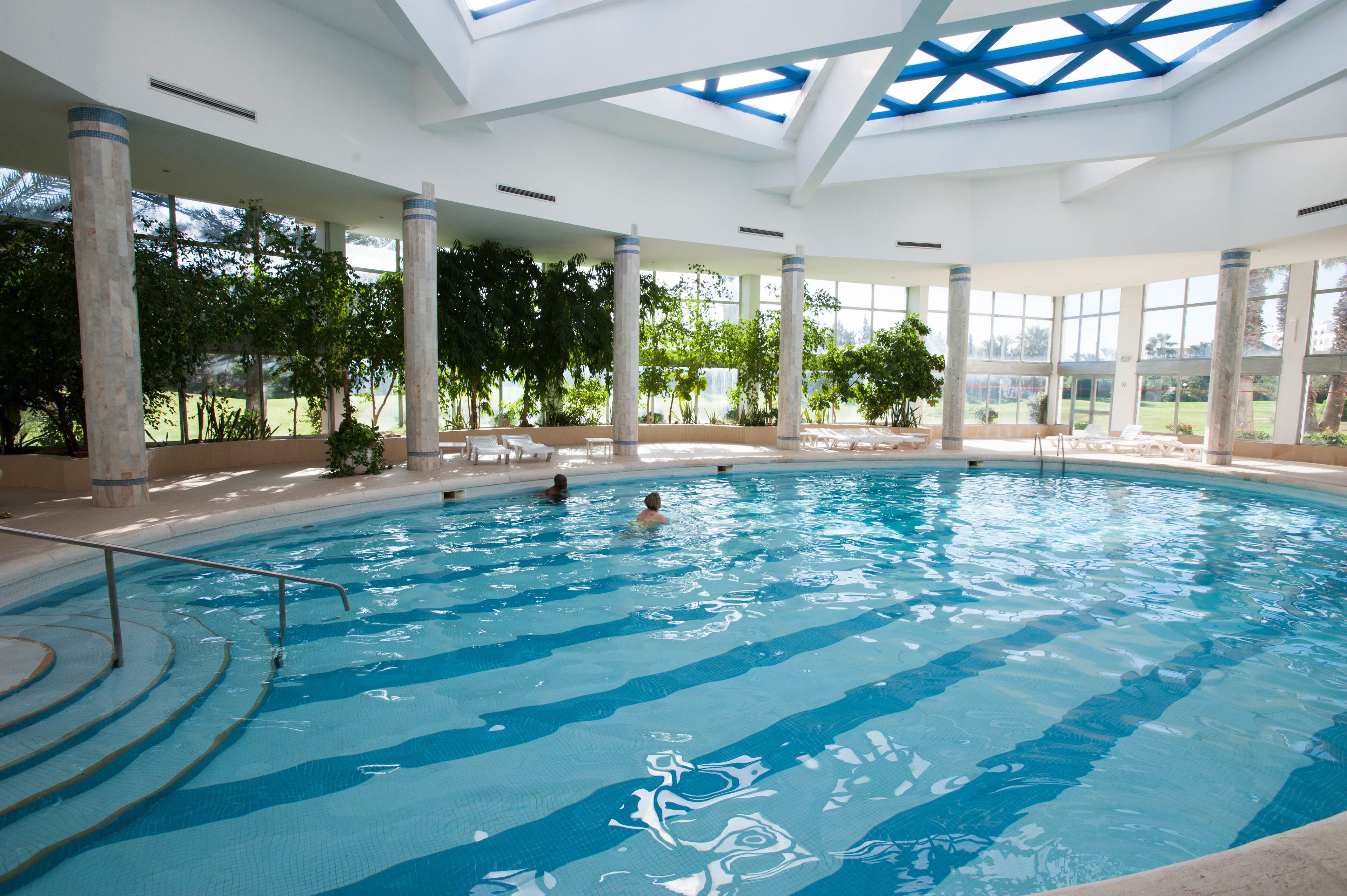 Une piscine intérieure à l’eau d’un bleu cristallin, entourée de grandes fenêtres et de colonnes.