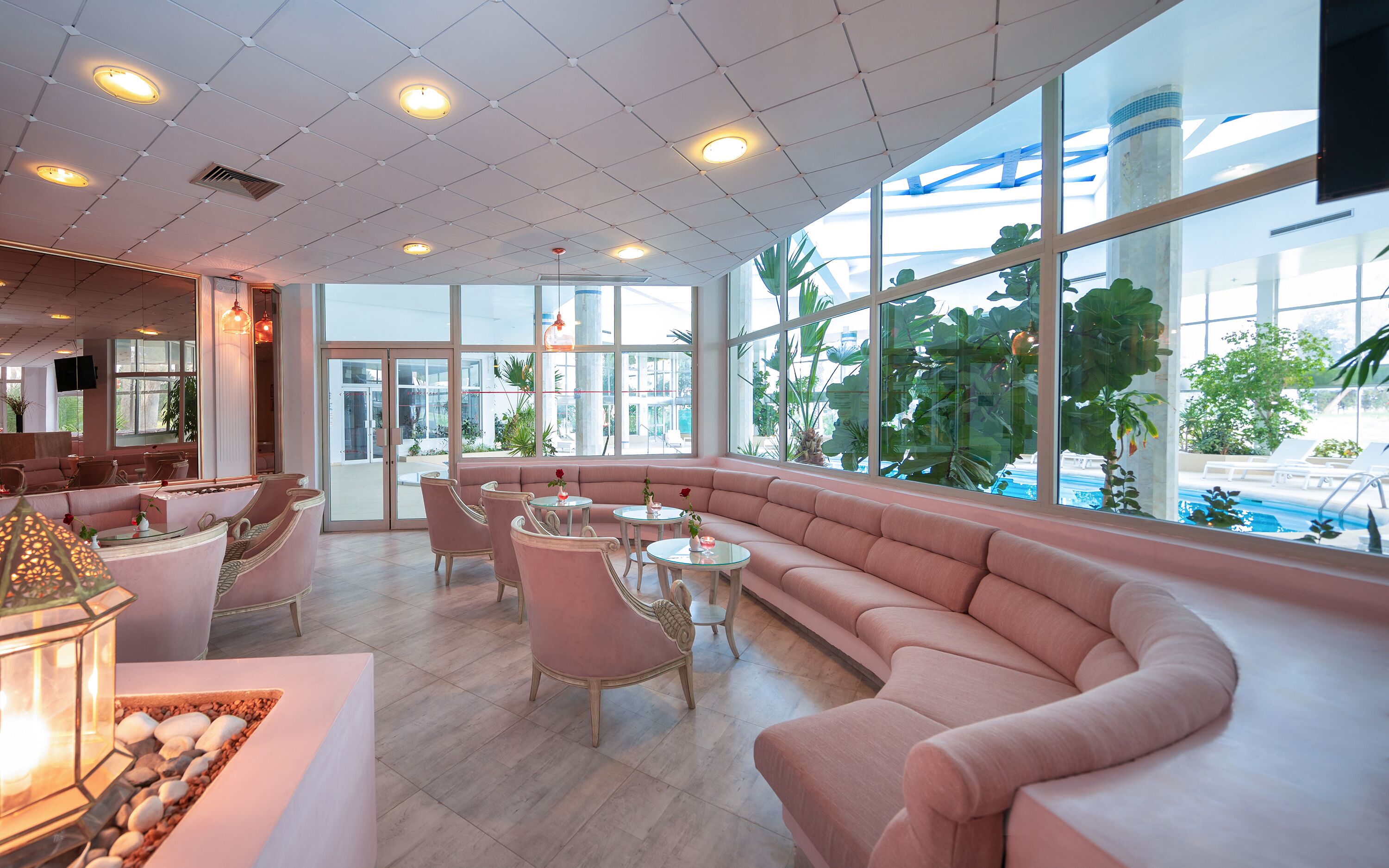 A spacious lounge with a curved pink sofa, round tables, and large windows offering a view of the outdoors.