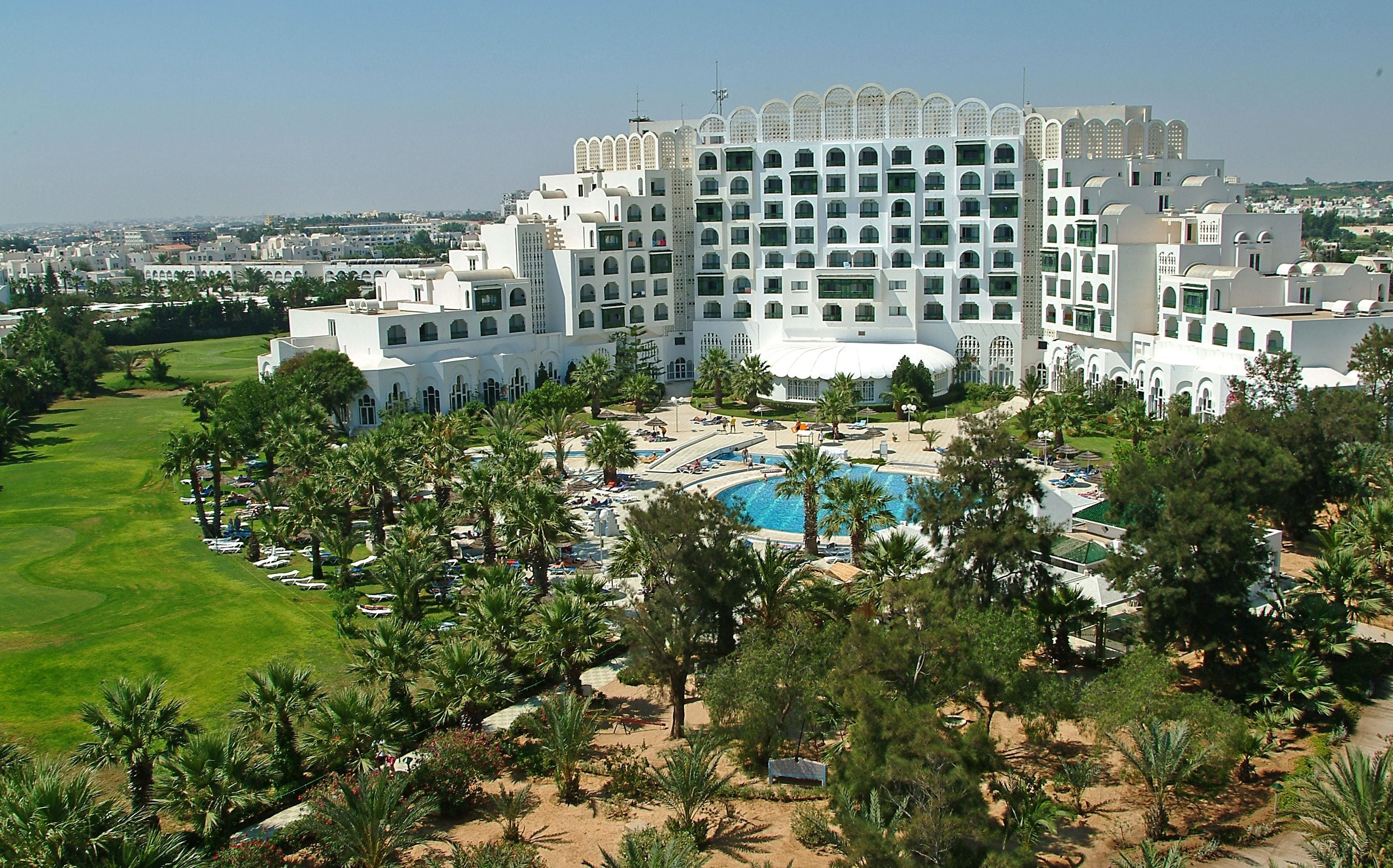 Un hotel grande con piscina central, rodeado de palmeras y un campo de golf.