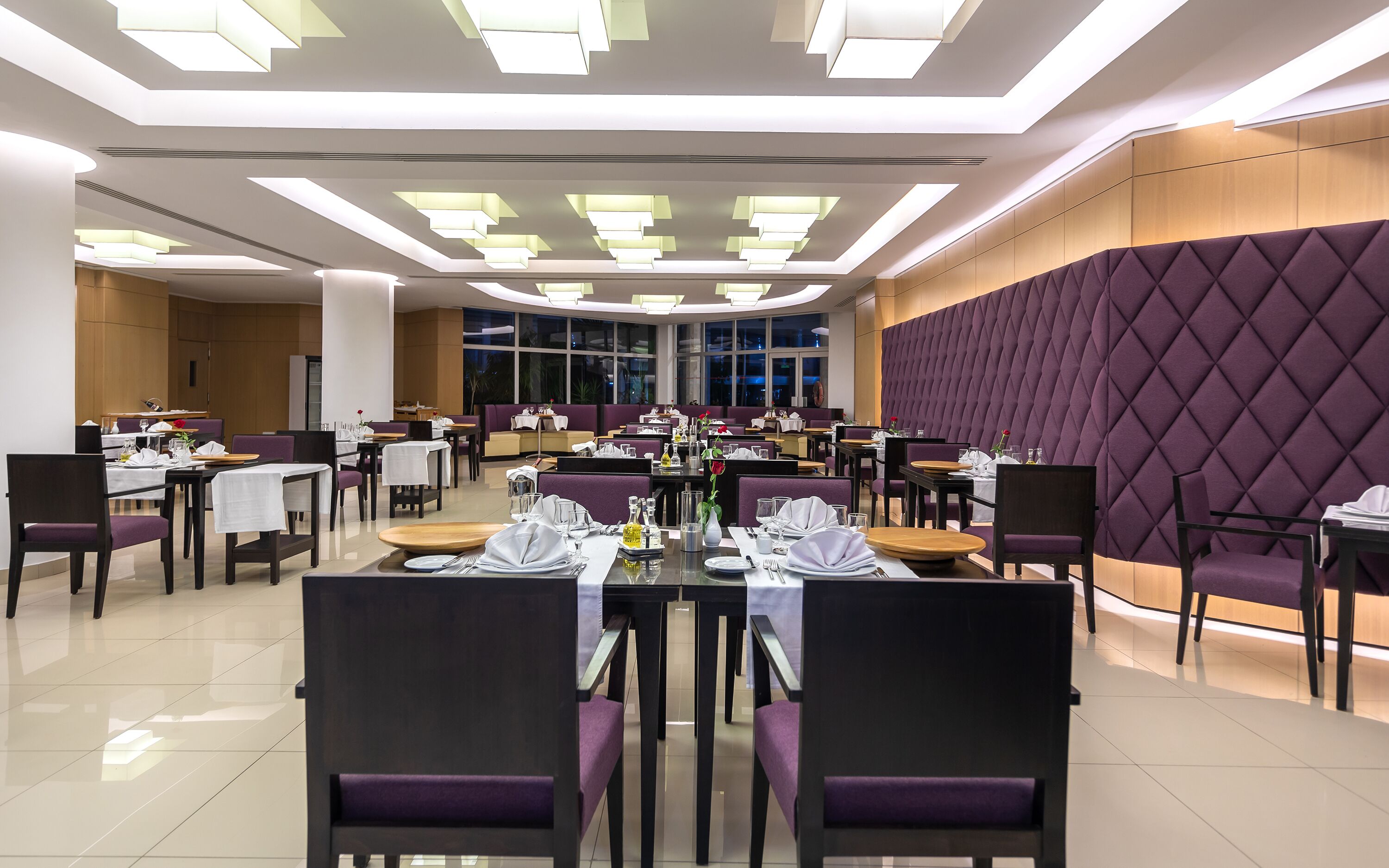 A modern dining area with tables set for service, a large window, and a high ceiling with recessed lighting.