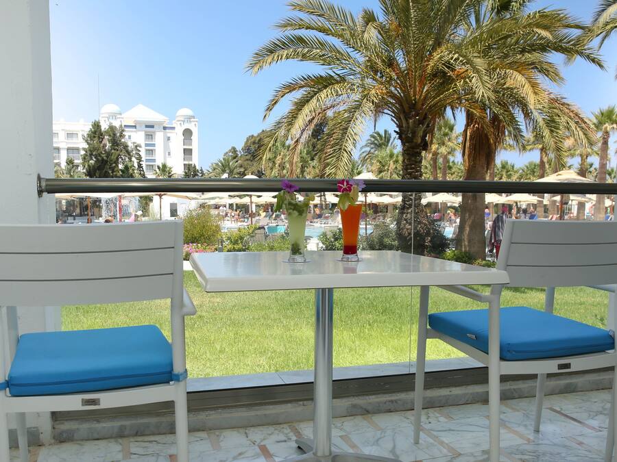 Varanda com duas cadeiras brancas e almofadas azuis, uma mesa pequena e vista para área da piscina com palmeiras e um edifício ao fundo.
