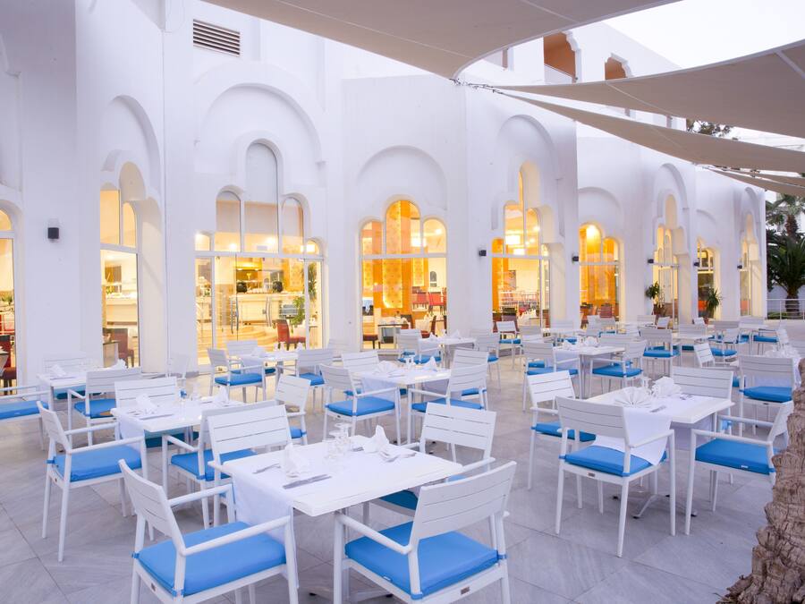 Un espace de restauration en plein air avec des tables blanches et des chaises bleues, entouré de murs blancs percés de fenêtres en arc de cercle.