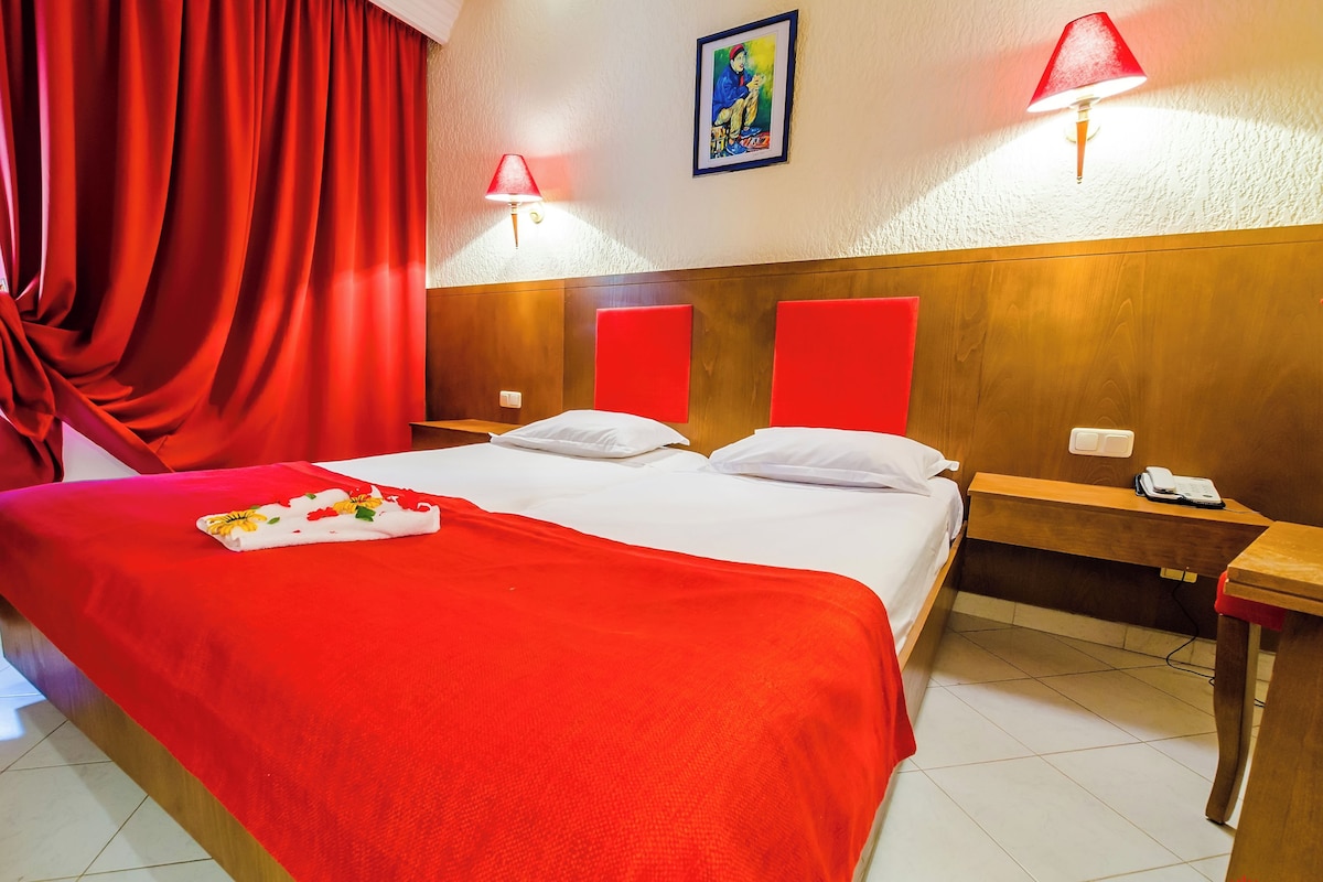A hotel room with a large bed, red curtains, two bedside lamps, a wooden headboard, and a painting on the wall.