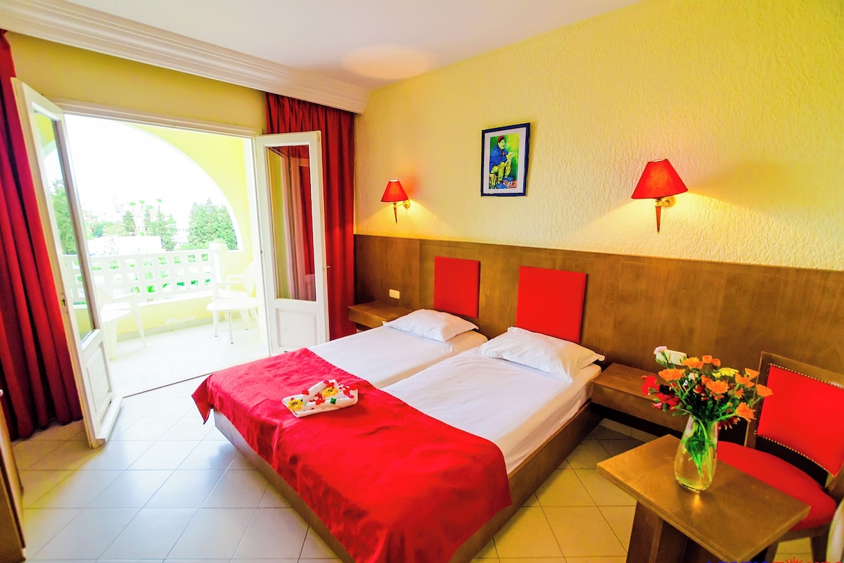 A hotel room with a bed, two bedside tables, a small table with a vase, a bench, and a door leading to an outdoor area.