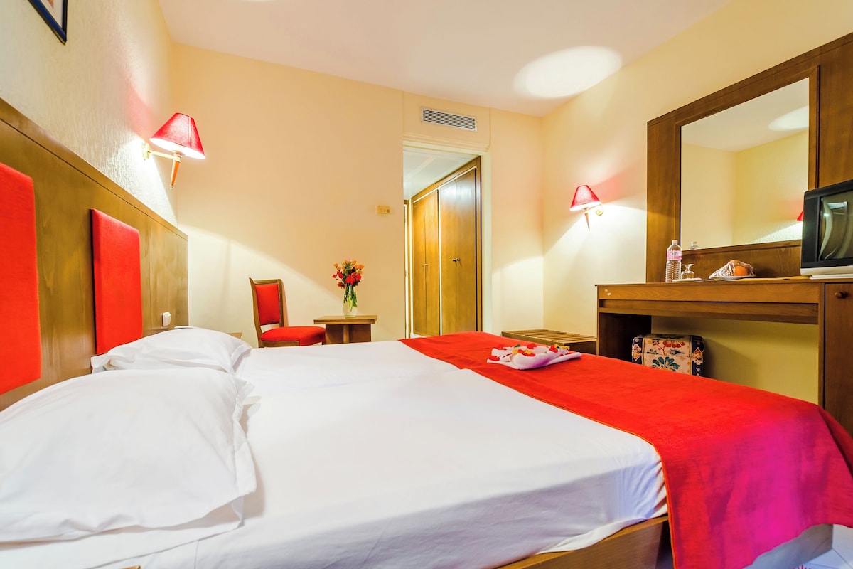 A hotel room with a bed, a chair, a small table with a vase, a mirror, and a television.