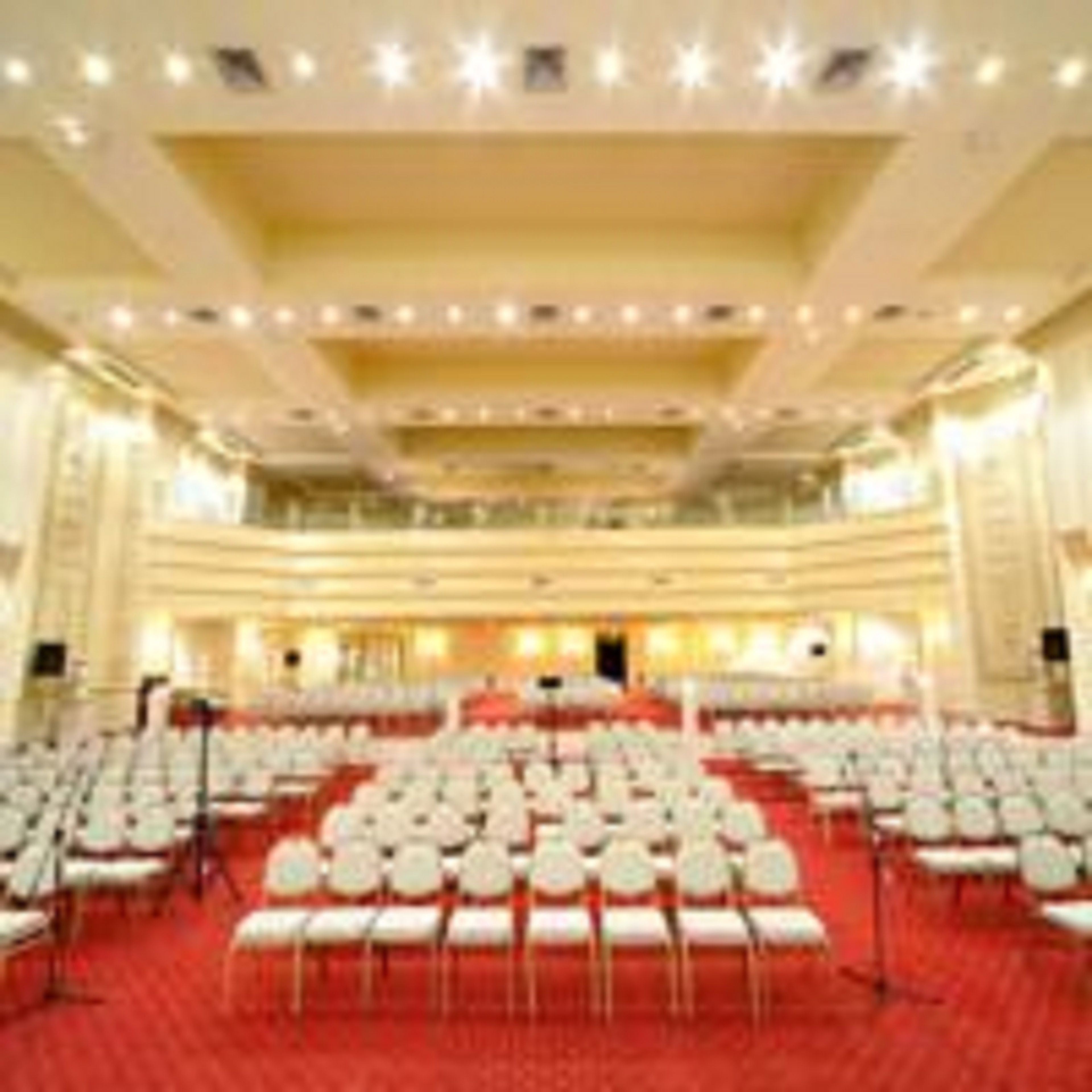A large conference room with rows of white chairs, a stage at the front, and a high ceiling with recessed lighting.