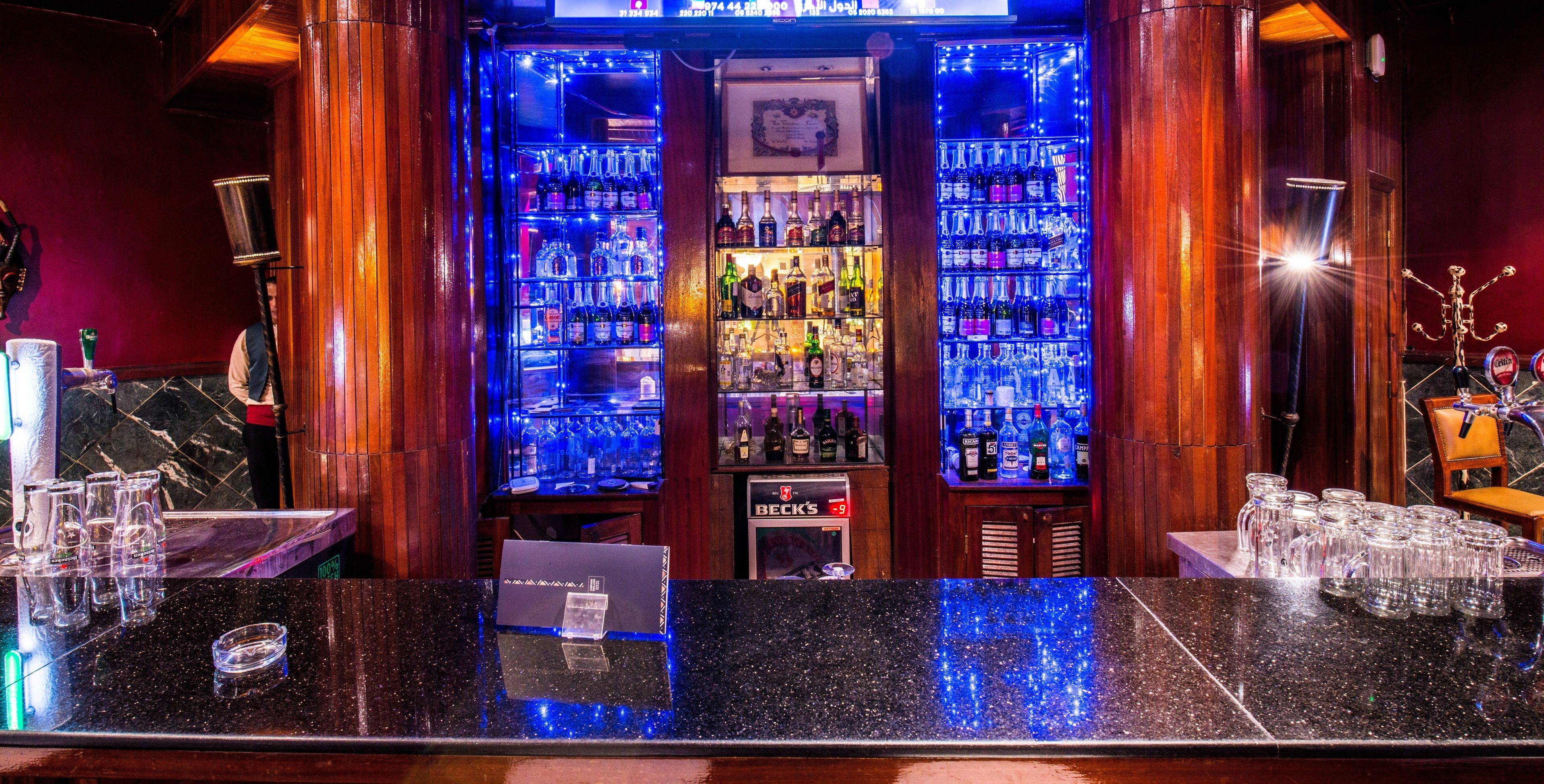 Un espace bar comprenant deux étagères éclairées où sont exposées des bouteilles et des verres, une caisse enregistreuse et un comptoir avec des verres et un menu.