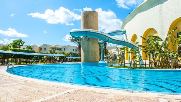 A swimming pool with a water slide, surrounded by palm trees and a building with yellow arches. - Carthage Thalasso Resort (La Marsa)