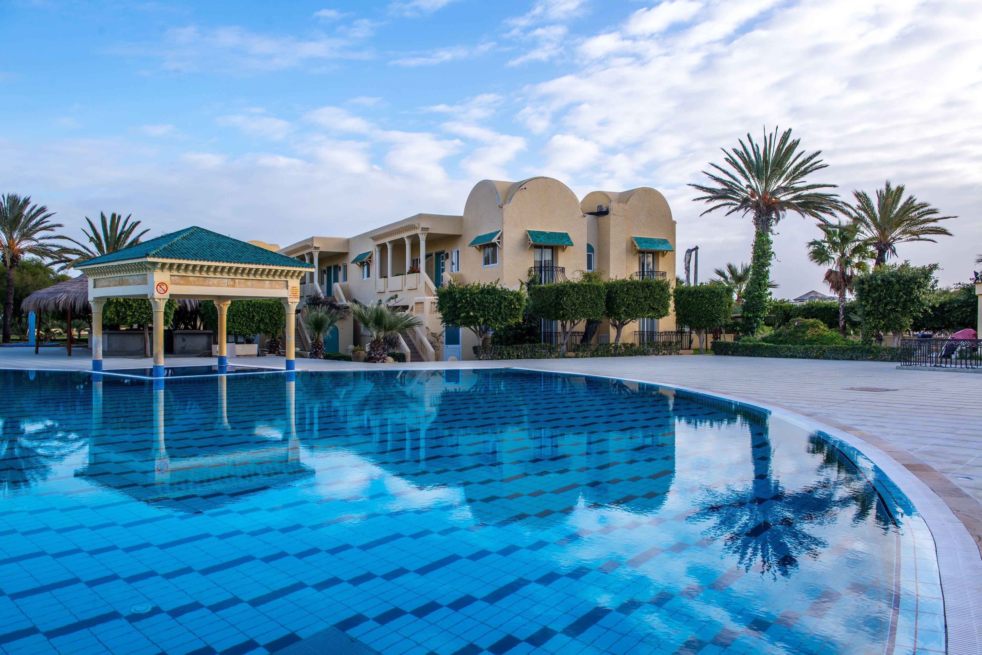 Uma piscina com deck revestido em azulejos, um gazebo e um edifício com janelas em arco e telhado verde.