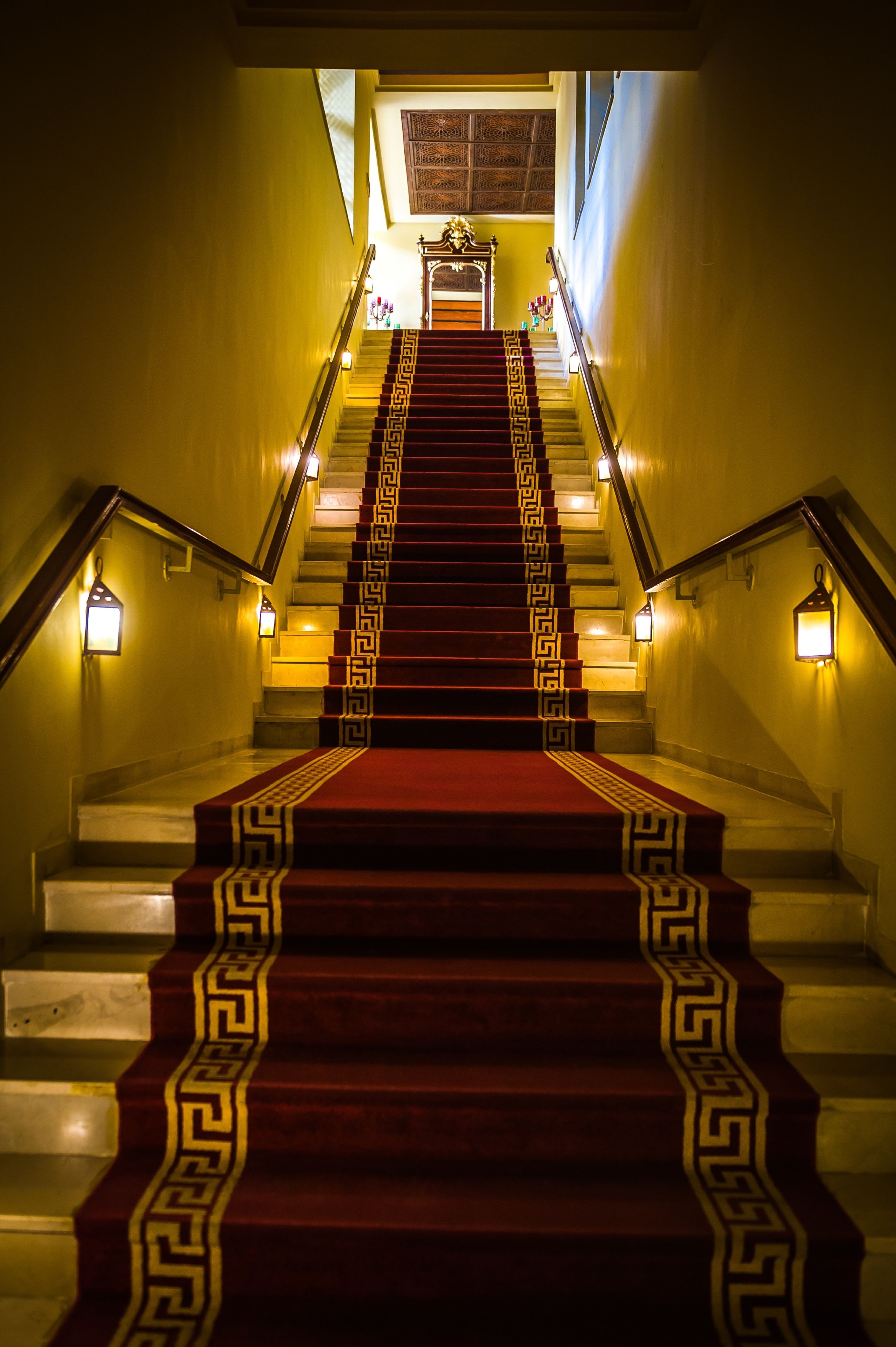 A grand staircase with a red carpet and decorative pattern, leading to a room with a patterned ceiling and wall sconces.